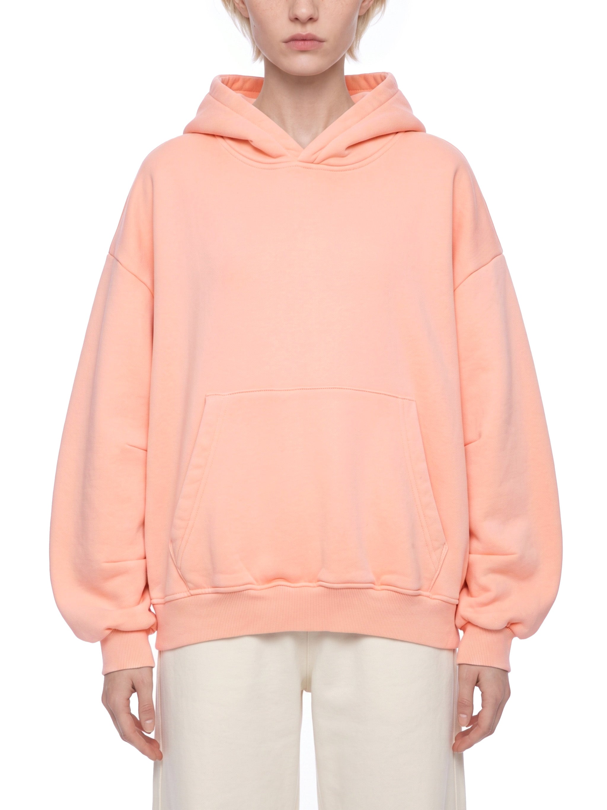 A model wearing an oversized peach-colored pullover hoodie with a kangaroo pocket, paired with cream-colored pants, shot against a solid white background.
