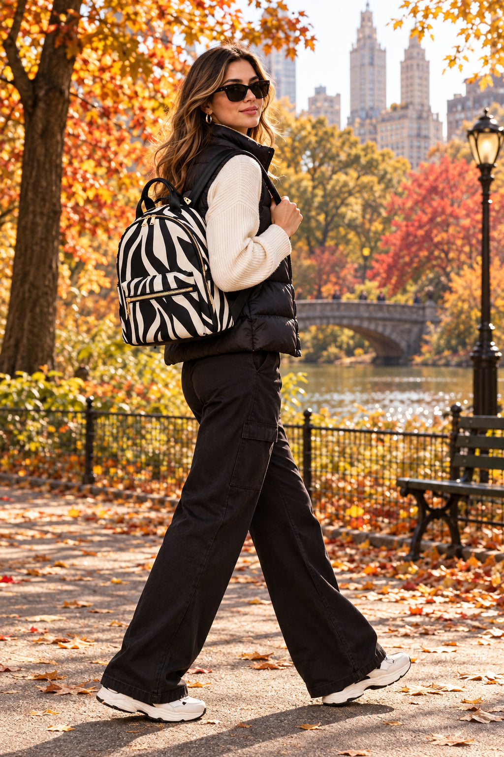 A zebra-print mini backpack with black and off-white stripes, featuring a top handle, a front zippered pocket with a gold zipper, and side pockets, shown against a light gray background.
